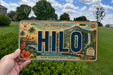 A person is holding a license plate with the word "HIlo" on it, which is a Hawaiian island. The license plate has a tropical landscape with palm trees, mountains, and a waterfall. The background of the image is a grassy field with trees.