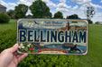 A person is holding a license plate that says "Bellingham" with a scenic background of mountains, buildings, and a body of water.