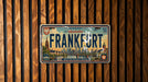 A vintage-style license plate from Germany that reads "FRANKFORT" is displayed against a wooden background.
