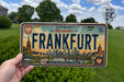 A person is holding a Germany license plate that says "FRANKFURT" in front of a cityscape.