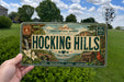 A person is holding a license plate with the text "HOCKING HILLS" and various scenic elements, such as a waterfall, bridge, and birds, against a background of trees and a blue sky.