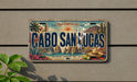 A vintage license plate with the text "CABO SAN LUCAS" printed on it, featuring a scenic illustration of a coastal town with palm trees, mountains, and a sunny sky.