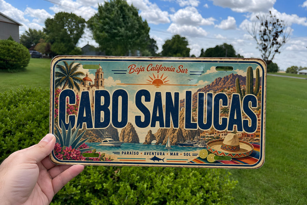 A person is holding a license plate that says "CABO SAN LUCAS" in bold letters. The license plate also features a scenic illustration of a coastal town with palm trees, mountains, and a body of water.