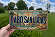 A person is holding a license plate that says "CABO SAN LUCAS" in bold letters. The license plate also features a scenic illustration of a coastal town with palm trees, mountains, and a body of water.