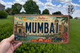 A vintage license plate from India, featuring the city of Mumbai, with a hand holding it in front of a backdrop of trees and a clear blue sky.