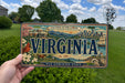 A person is holding a Virginia license plate with a scenic illustration of the state, including mountains, buildings, and a bird.
