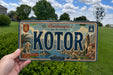 A person is holding a vintage license plate from the town of Kotor, which features a scenic illustration of the town and its surroundings.