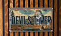 A license plate with the text "Devil Tower" and various scenic elements, including a mountain, trees, and a person on horseback, all set against a wooden background.