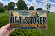 A person is holding a license plate that features the text "Devils Tower" and "Northeast Wyoming", with a scenic illustration of the Devils Tower and surrounding landscape.