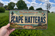 A person is holding a license plate that says "Cape Hatteras" with a lighthouse and beach scene on it.