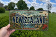 A person is holding a New Zealand license plate in front of a scenic landscape with mountains, a lighthouse, and a body of water.