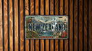 A vintage license plate from New Zealand, featuring a scenic landscape with mountains, a lighthouse, and a boat, is displayed on a wooden wall.