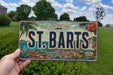 A person is holding a license plate that says "ST. BARTS" against a background of a tropical island landscape with palm trees, flowers, and a body of water.