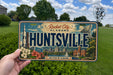 A person is holding a license plate that says "HUNTSVILLE" with a cityscape and rocket ship illustration on it.