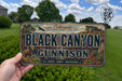 A person is holding a license plate that says "Black Canyon of the Gunnison" with a scenic background of trees and a house.