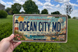 A person is holding a license plate with a beach scene and a lighthouse, against a backdrop of trees and a blue sky.
