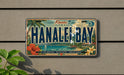 A vintage license plate from Hawaii, featuring the text "Kauai Hanahe Bay" and surrounded by tropical imagery such as palm trees, flowers, and a beach.