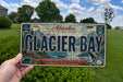 A person is holding a license plate that says "Glacier Bay" in front of a scenic landscape with mountains and a body of water.