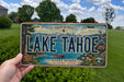 A person is holding a license plate with the text "LAKE TAHOE" and "SIERRA NEVADA CALIFORNIA, NEVADA" printed on it. The license plate also features a scenic image of Lake Tahoe and mountains, as well as various stickers and text related to the location.