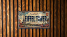 A vintage license plate from Paris, with the Eiffel Tower and other Parisian landmarks visible, is displayed on a wooden wall.
