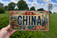 A person is holding a license plate with the word "CHINA" prominently displayed on it. The license plate features an illustration of various landmarks and scenes from China, including a temple, pagodas, and a river.