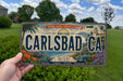 A person is holding a license plate that says "CARLSBAD CA" against a background of a grassy field and trees.