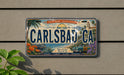 A vintage license plate fromCARLSBAD CA, featuring a beach scene and a car, is displayed on a wall.