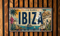 A vintage license plate from the island of Ibiza, featuring a tropical landscape with palm trees, cacti, and other scenic elements.