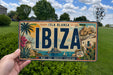 A person is holding a license plate with the text "IBAZ" on it, which is a tropical island in the Mediterranean.