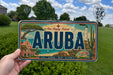 A person is holding a license plate with the word "ARUBA" prominently displayed, which is the name of the island. The license plate also features illustrations of various landmarks and scenery, including a lighthouse, palm trees, and a beach.