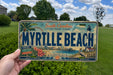A person is holding a license plate that says "MYRTILLE BEACH" with a scenic beach and lighthouse image on it.