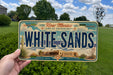 A person is holding a New Mexico license plate that says "WHITE SAND".