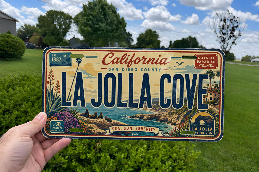 A person is holding a California license plate that reads "LA Jolla Cove" against a backdrop of a grassy field and trees.