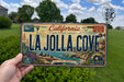 A person is holding a California license plate that reads "LA Jolla Cove" against a backdrop of a grassy field and trees.