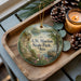 A wooden tray with a decorative ornament featuring a scenic landscape, including a lake, mountains, and pine cones, along with a lit candle and pine cones.