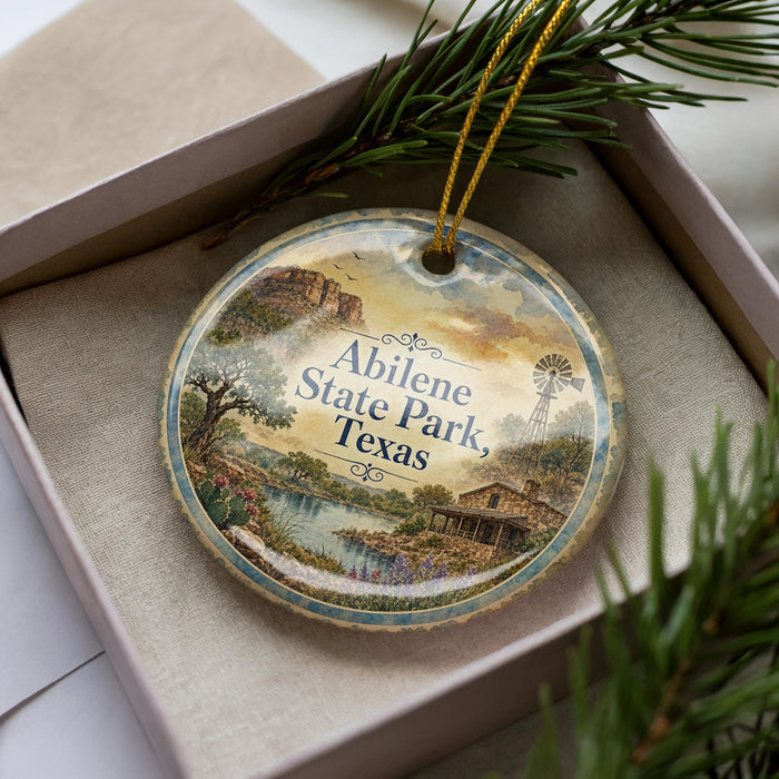 A decorative ornament in a box, featuring a scenic landscape with a river, windmill, and trees, along with the text "Abilene State Park, Texas".