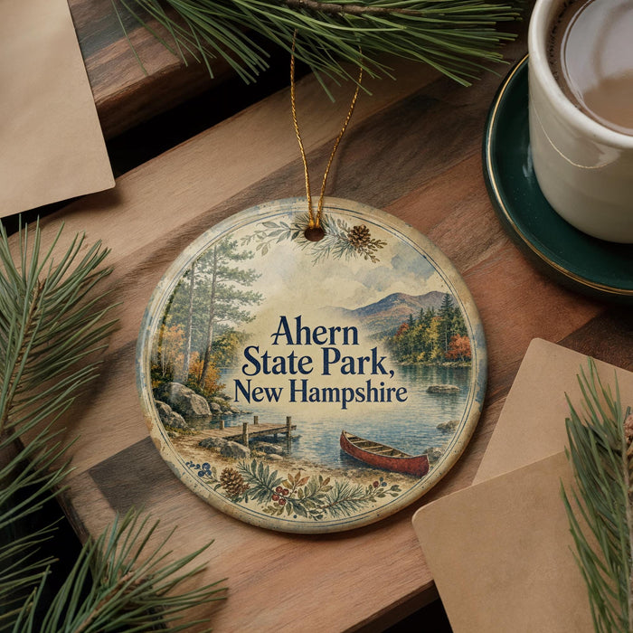 A decorative ornament with a scenic image of a lake and mountains, featuring a canoe, is displayed on a wooden surface alongside a cup of coffee and some pine branches.