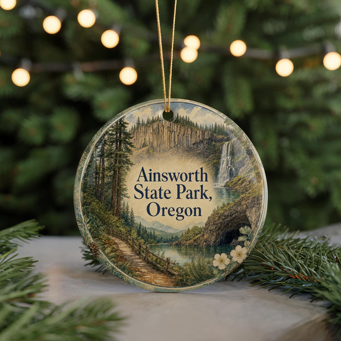 A decorative ornament featuring a scenic landscape with a waterfall, trees, and a path, hanging in front of a Christmas tree with lights.