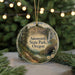 A decorative ornament featuring a scenic landscape with a waterfall, trees, and a path, hanging in front of a Christmas tree with lights.