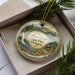 A decorative ornament in a box, featuring a scenic landscape with a waterfall, pine trees, and a cabin.