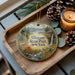 A decorative ornament featuring a scenic landscape of Allegany State Park, New York, with a deer and a waterfall, placed on a wooden tray alongside pine cones and a lit candle.
