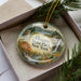 A decorative ornament in a box, featuring a scenic landscape with a deer, trees, and a waterfall.