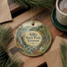 A round ornament with a scenic landscape of Allis State Park, Vermont, featuring a waterfall and pine trees, is displayed on a wooden surface alongside a cup of coffee and some papers.
