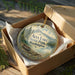 A round ornament in a wooden box, featuring a scenic landscape with mountains, trees, and a lake, with the text "Alta Lake State Park, Washington" displayed on it.