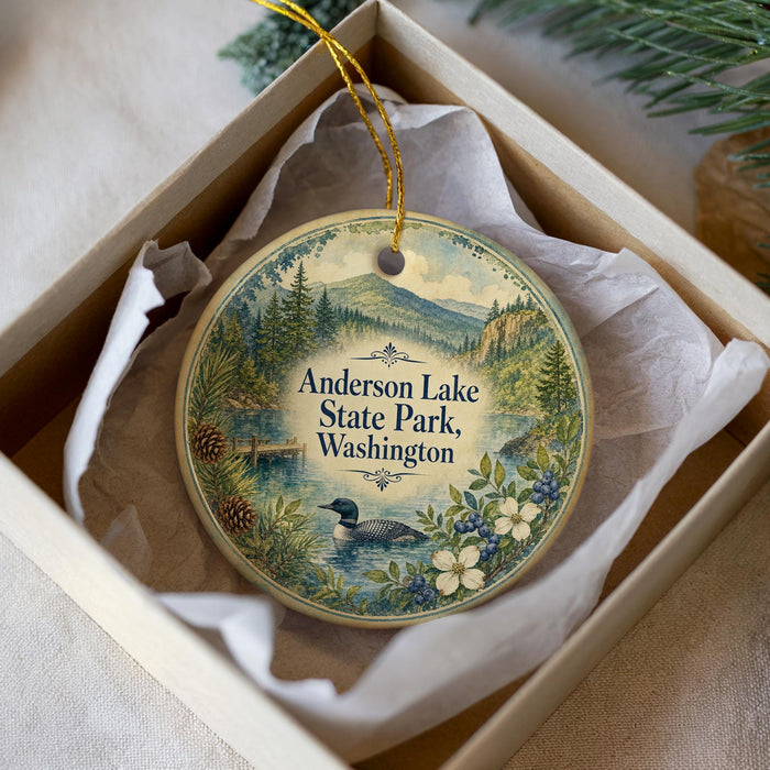 A decorative ornament in a white box, featuring a landscape scene with a lake, mountains, pine trees, and a bird.