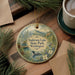 A decorative ornament with a lake and forest scene, featuring a bird and pine trees, is displayed on a wooden surface alongside a cup of coffee and some papers.