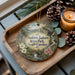 A decorative ornament with a scenic landscape of Andrew Jackson State Park, South Carolina, featuring a bridge, pine trees, and flowers. The ornament is displayed on a wooden tray, accompanied by a lit candle and pine cones.