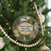 A round ornament hanging on a Christmas tree, featuring a scenic landscape with a bridge and flowers.
