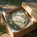 A decorative ornament in a wooden box, featuring a scenic landscape with a river and a house, is placed on a wooden surface.