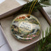 A decorative ornament in a box, featuring a scenic landscape with a waterfall, pine trees, and a barn.